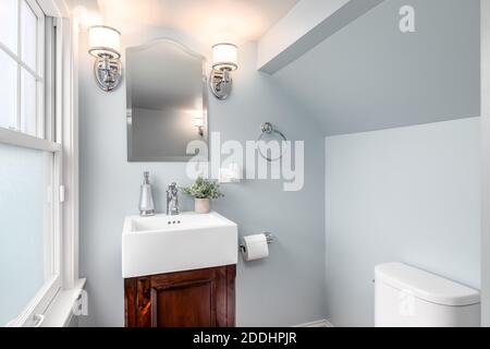 Eine kleine Toilette mit blauen Wänden, einem Holzwaschtisch, Blumen auf dem weißen Waschbecken und Lampen neben dem Spiegel. Stockfoto