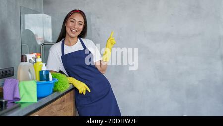 Fröhliche junge Frau, Putzfrau in Schutzhandschuhen lächelnd auf Kamera, zeigt auf, während in der Küche mit Reinigungsprodukten und Ausrüstung, bereit für die Reinigung des Hauses stehen. Ordnung Und Sauberkeit Stockfoto