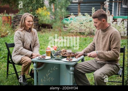 Ein Mann legt Teller mit gebratenem Fleisch auf den Picknicktisch. Die letzten warmen Herbsttage vor dem Winter. Gemütliches Picknick im Hinterhof eines privaten Hauses Stockfoto