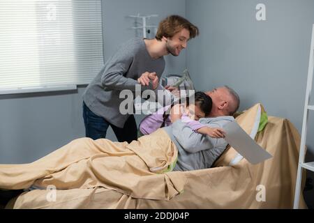 Enkel, ein kleines Mädchen und ihr älterer Bruder besuchen ihren Opa in einer Krankenstation, das kleine Mädchen umarmt ihren Opa und der ältere Junge schüttelt seine Hand. Stockfoto