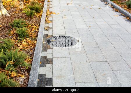 Schachtabdeckung rund schwarz Metallgitter auf der Gasse Stadt park gepflastert mit Granit grauen Fliesen mit einem Steinrand Nahaufnahme Herbstsaison mit Herbst Stockfoto