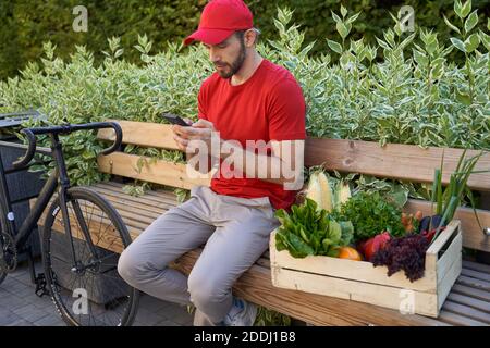 App. Lebensmittelzustellung Männlicher Kurier in Uniform sitzt auf der Bank im Freien und mit dem Handy, während Lebensmittel-Box mit frischem Gemüse auf dem Fahrrad zu liefern Stockfoto