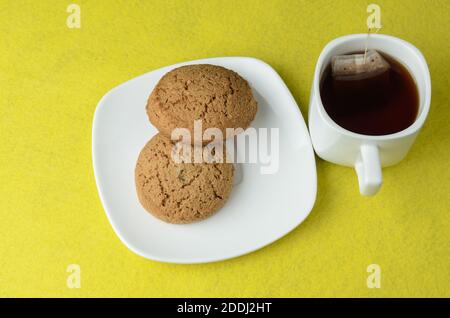 Haferflocken-Kekse auf einem weißen Teller mit einer Tasse Tee auf einem verschwommenen gelben Hintergrund. Selektiver Fokus. Stockfoto