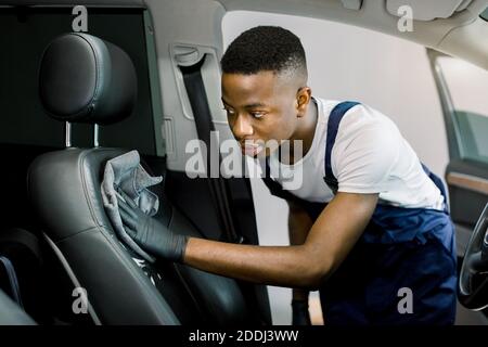 Auto Detaillierung und Valeting Konzept. Schöner afroamerikanischer Mann Autodienstmitarbeiter, trägt weißes T-Shirt und blaue Overalls, Reinigung Auto Innenraum Stockfoto