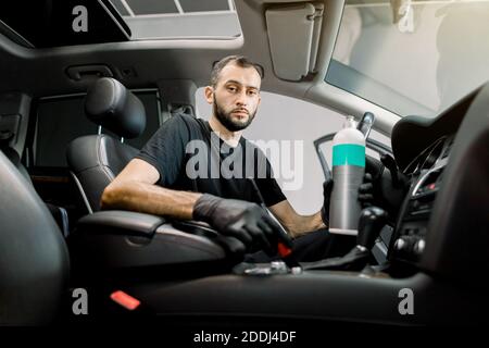 Junge konzentrierte männliche Auto-Service-Arbeiter Reinigung Staub aus dem Inneren des Luxusautos, mit speziellen Bürste und Kunststoff Pflege professionelles Produkt in Stockfoto