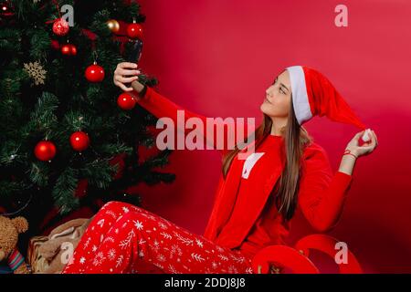 Ein Mädchen in Santa hat macht ein Selfie auf einem Schlitten, zu Hause bei einem Weihnachtsbaum. Stockfoto