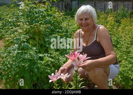 Lächelnd im Alter blonde Frau in Camisole oben ist hunker unten in der Nähe rosa Lilien und Himbeerpflanzen im Gemüsegarten auf dem ländlichen Sommerhof. Stockfoto