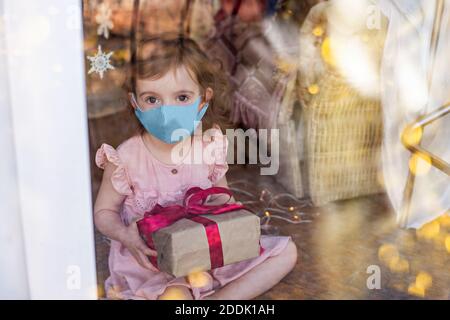 Kleines schönes Mädchen in einer medizinischen Maske ist traurig am Fenster des Hauses, hält ein Neujahrsgeschenk in ihren Händen. Das Baby wird nicht bekommen, einen Wunsch zu machen Stockfoto