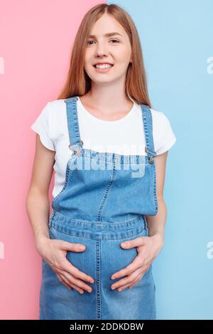 Eine schöne junge schwangere emotionale Frau in einem Jeansjumpsuit Posen auf einem isolierten rosa und blauen Hintergrund Stockfoto