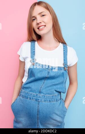 Eine schöne junge schwangere emotionale Frau in einem Jeansjumpsuit Posen auf einem isolierten rosa und blauen Hintergrund Stockfoto