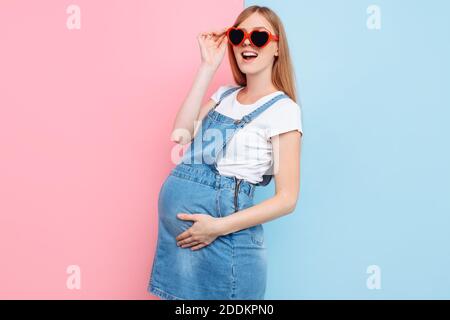 Emotional glücklich junge schwangere Frau in Denim Overalls, trägt herzförmige Brille, posiert auf einem isolierten rosa und blauen Hintergrund Stockfoto