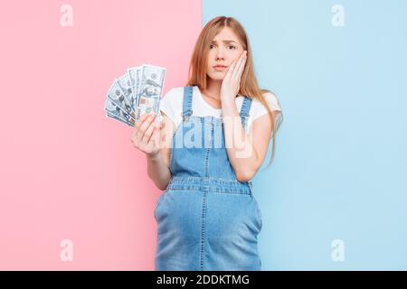 Glückliche junge schwangere Frau hält Dollarscheine, Mädchen, auf einem rosa und blauen Hintergrund, das Konzept der Leihmutterschaft stehen Stockfoto