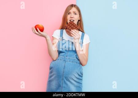 Eine perplex schwangere Frau wählt zwischen Süßigkeiten und Obst, hält einen Apfel und Schokolade, stehen auf einem rosa und blauen Hintergrund Stockfoto