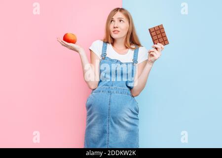 Eine perplex schwangere Frau wählt zwischen Süßigkeiten und Obst, hält einen Apfel und Schokolade, stehen auf einem rosa und blauen Hintergrund Stockfoto