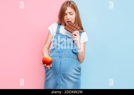 Eine perplex schwangere Frau wählt zwischen Süßigkeiten und Obst, hält einen Apfel und Schokolade, stehen auf einem rosa und blauen Hintergrund Stockfoto