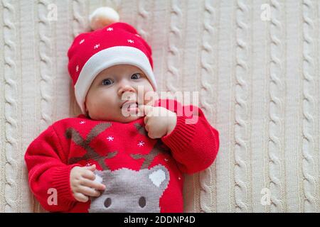 Nahaufnahme Porträt des Neugeborenen. Niedliche kaukasische Baby Mädchen 3-4 Monate alt in Santa Kostüm liegt auf gestrickte gemütliche Decke auf dem Bett. Frohe Weihnachten Weihnachten Stockfoto