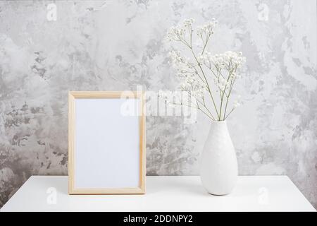 Holz vertikal weißen leeren Rahmen und Strauß von weißen kleinen Blumen Gypsophila in Vase auf Tisch auf grauen Beton Wand Hintergrund. Mockup-Vorlage für Stockfoto