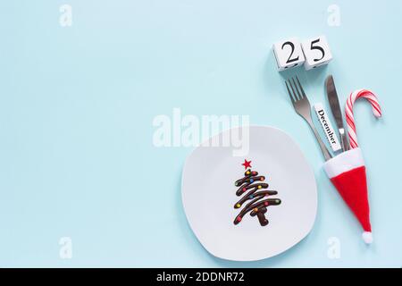 Weihnachtskomposition Kalender Dezember 25. Weihnachtsbaum mit süßer Schokolade auf Teller, Besteck in weihnachtsmütze auf blauem Hintergrund. Flat Lay Draufsicht Kopie en Stockfoto