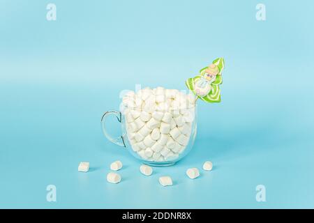 Große transparente Tasse Marshmallows mit grünen Lollipop weihnachtsbaum auf blauem Hintergrund. Frohe Weihnachten oder Happy New Year Konzept. Minimalistischer Stil, T Stockfoto