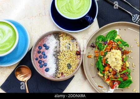 Smoothie Schüssel mit rohen Lebensmitteln und Toast mit Hummus und gebackenem Gemüse und Matcha Latte Tasse grünen Tee auf dem Tisch Stockfoto