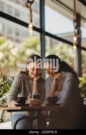 Freunde, die in einem Café sitzen und auf ein Smartphone blicken und lächeln. Zwei Frauen treffen sich in einem Kaffee mit einem Handy. Stockfoto