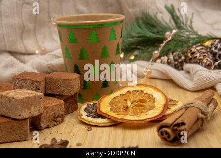 Einweg-Weihnachtsglas aus Pappe mit einem Getränk und verschiedenen Süßigkeiten und aromatischen Gewürzen auf einem Holztablett. Stockfoto