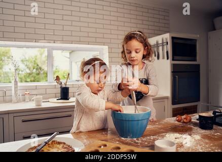 Kleine Schwestern genießen es, Teig mit Eiern zum Backen zu machen Moderne, verworaufene Küche Stockfoto