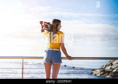 Eine junge kaukasische Frau mit blonden Haaren, die ein Skateboard hält Und genießen Sie den Meerblick Stockfoto