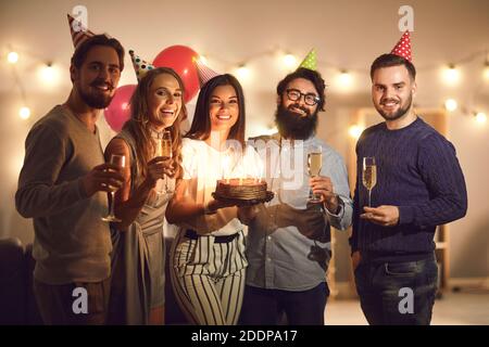 Junge lächelnde Freunde in Weihnachtsmützen stehen mit Geburtstagstorte Und Champagner Stockfoto