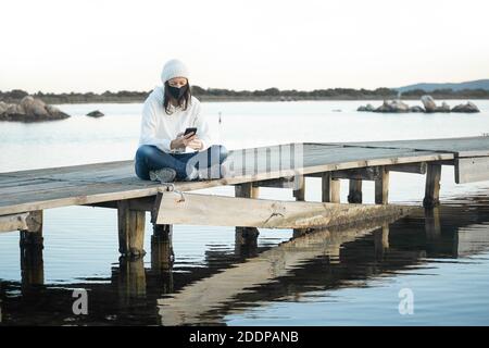 Einsame Frau lebt Natur sitzt auf einem Pier bei Sonnenuntergang Mit Smartphone zu chatten trägt Coronavirus sicheren Schutz medizinische Maske - Neue communicatio Stockfoto