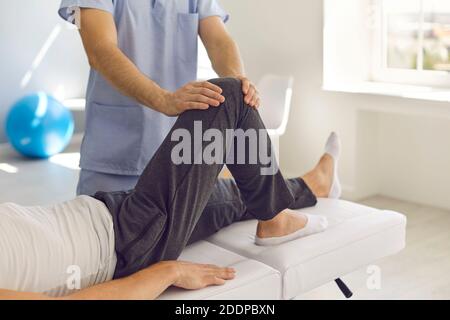 Der abgeschnittene Schuss des Physiotherapeuten führt eine medizinische Behandlung am Bein des Patienten durch Massage seines Knies durch. Stockfoto