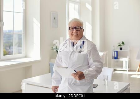 Erfahrener Oberarzt steht im Krankenhausbüro mit Klemmbrett in Hand auf die Kamera schauen Stockfoto