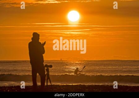 Swansea, Großbritannien. November 2020. Ein Fotograf reibt sich die Hände zusammen, während er einen Surf-Skifahrer sieht, der das Beste aus dem Wellengang macht, denn der Ozean ist heute Morgen in der Langland Bay bei Swansea im goldenen Glanz des Sonnenaufgangs gebadet. Quelle: Phil Rees/Alamy Live News Stockfoto