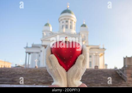 In den Händen eines Christbaumspielzeugs in Form eines Herzens auf dem Hintergrund des berühmten Himmelfahrtsamts in Helsinki. Konzept Ich liebe Helsinki, Frohes neues Jahr. Hochwertige Fotos Stockfoto