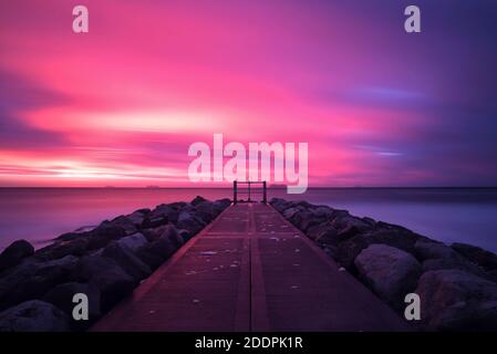 Eine lange Belichtung fängt einen farbenfrohen Sonnenaufgang ein, während Wolken durch einen rosa und violetten Himmel Streifen. Der Steinweg auf einer Groyne führt hinaus ins Meer. Stockfoto