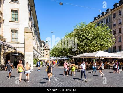 Leute, die spazieren gehen oder einen Drink in einem Straßencafé auf dem Molard Platz in Genf nehmen, das an einem sonnigen Tag vom Molard Turm überblickt wird. Stockfoto