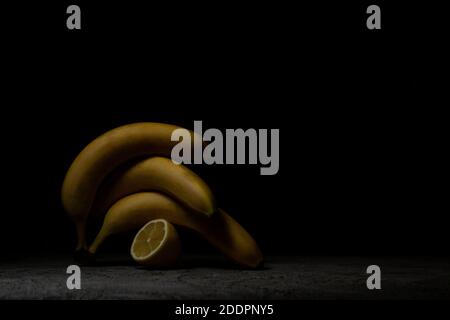 Drei reife Bananen liegen übereinander auf dunkelgrauem Beton, neben einer Zitrone. Low-Taste. Stockfoto