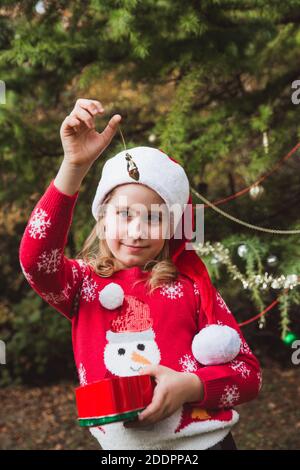 Frohe Weihnachten und frohe Feiertage. Kleines Mädchen in roten Pullover und weihnachtshut Dekoration der Weihnachtsbaum im Freien im Hof des Hauses vor Stockfoto