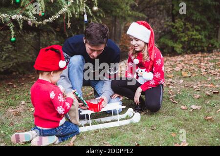 Vater und zwei Töchter öffnen Geschenke Boxen in der Nähe Weihnachtsbaum im Freien im Hof des Hauses. Frohe Weihnachten und frohe Feiertage Stockfoto