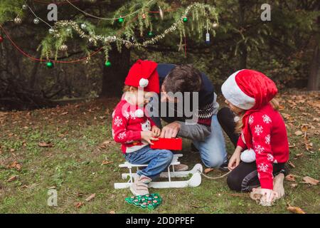 Vater und zwei Töchter öffnen Geschenke Boxen in der Nähe Weihnachtsbaum im Freien im Hof des Hauses. Frohe Weihnachten und frohe Feiertage Stockfoto