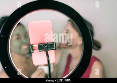 Mama und Tochter lächeln süß und Anwendung Maske auf ihr Gesicht und suchen das Smartphone. Heiter schöne Freunde nehmen Selfie zu Hause mit Gesichts c Stockfoto