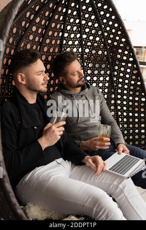 Junge gutaussehende Homosexuell Paar entspannen auf der Schaukel auf dem Balkon mit Bier und Browsen Social Media auf Laptop. Hochwertige Fotos Stockfoto