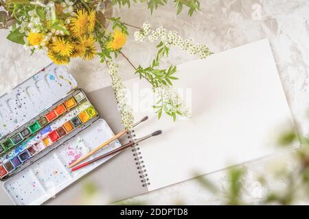 Draufsicht auf einen Tisch mit einem offenen Skizzenbuch, Farben und Pinsel neben einem Blumenstrauß, Kopierraum. Freizeit Stockfoto