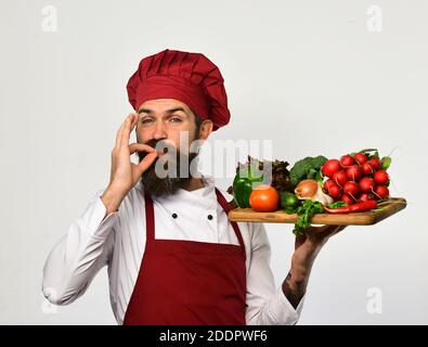 Kochen Sie mit zufriedenen Gesicht in burgunderroten Uniform hält Salat Zutaten. Kochen und vegetarische Ernährung Konzept. Mann mit Bart isoliert auf weißem Hintergrund. Koch zeigt Super Tasty Schild mit Gemüse Stockfoto