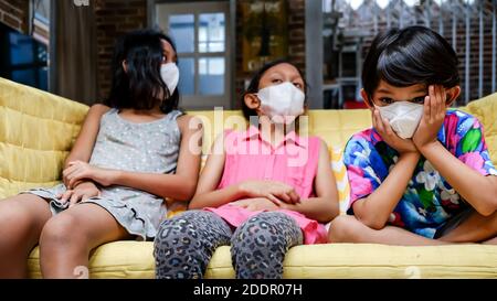 Südostasiatische Kinder tragen medizinische Gesichtsmaske auf Sofa sitzen Zusammen Fühlen Sich Während Der Isolation Zu Hause Gelangweilt Stockfoto