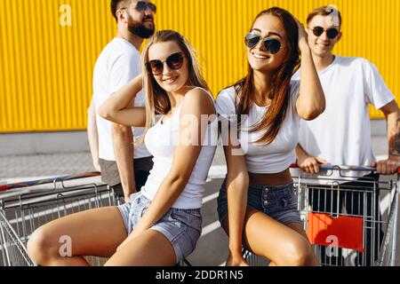 Eine Gruppe von glücklichen jungen Menschen, Männer und Frauen, die Spaß auf Trolleys in der Nähe eines Supermarkts, posiert auf einem Trolley an einem Sommertag mit Sonnenschein. Stockfoto