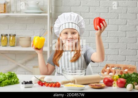 Das Kind hält gelbe und rote Paprika. Kleines Mädchen in Chefhut und eine Schürze Kochen Pizza in der Küche. Stockfoto