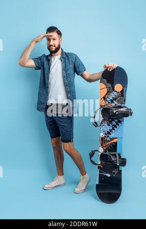 Ein glücklicher bärtiger Mann, in Shorts und Hemd gekleidet, hält ein Snowboard und blickt in die Ferne, steht auf einem isolierten blauen Hintergrund Stockfoto