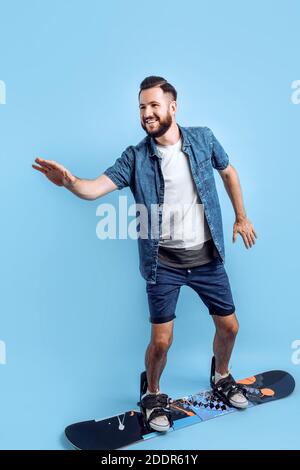 Extremer Mann, der auf einem isolierten blauen Hintergrund steht und mit seinen Füßen auf einem Snowboard in die Ferne blickt Stockfoto