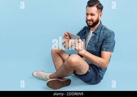 Ein attraktiver bärtiger Mann, in stilvoller Freizeitkleidung, hält ein Smartphone in den Händen und schaut auf den Telefonbildschirm, der auf blauem Hintergrund sitzt Stockfoto